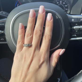 Sassy Nails - Nails - A close-up of a person's left hand resting on a car's steering wheel. The hand has well-manicured nails painted in a light pink or nude color and is wearing a silver ring with multiple stones. The background shows part of the car's dashboard and instrument panel, indicating the photo was taken inside a vehicle. - Nail Salons Near Me - Schaumburg, Illinois