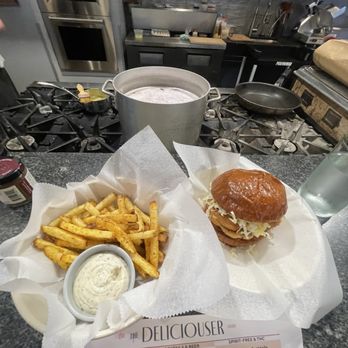 The Deliciouser - Best chicken sandwich ever! - A burger with shredded cheese and a bun is served on a paper-lined tray alongside seasoned French fries and a small container of dipping sauce, placed on a kitchen countertop. The background shows a professional kitchen with a stove, knives, and kitchen utensils, indicating a cooking or dining environment. - Herbs & Spices Near Me - Madison, Wisconsin