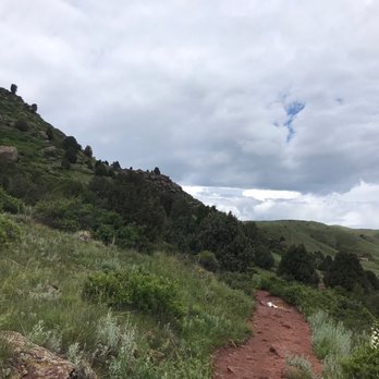 Red Rocks Park - A dirt hiking trail runs through a lush green hillside with trees and shrubs, under a cloudy sky, in a natural outdoor setting. - Parks Near Me - Morrison, Colorado
