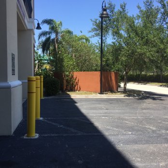 CubeSmart Self Storage - non-gated parking lot - A parking lot adjacent to a building features yellow safety posts, trees, and a street lamp, with a clear blue sky overhead. - Self Storage Near Me - Miramar, Florida