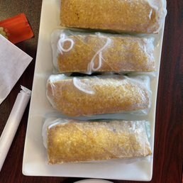 Spring Rolls - Four Vietnamese spring rolls are placed on a white rectangular plate, each wrapped in clear plastic. The rolls have a golden-brown exterior, indicating they are fried. Some white sauce or decorative drizzle is visible on the wrappers. The plate is on a dark wooden table, with a napkin, chopsticks, and a small red packet nearby.