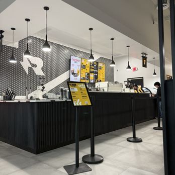 Refuel Coffee - Where to order - The image shows a coffee shop counter with a black, vertically grooved surface. Above the counter, there are multiple black pendant lights hanging from the ceiling. Behind the counter, digital menu boards display drink options, and a patterned wall with a large letter 'R' and star design is visible. The floor is tiled in light gray, and there are a few customers and staff members present. - Coffee & Tea Near Me - St. Augustine, Florida