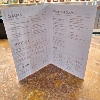 Solette - A printed menu on a table displays drink options, with a bar featuring numerous liquor bottles in the background. - American Near Me - Chicago, Illinois