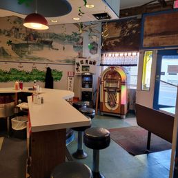 Dining area - The image shows the interior of a diner with a bar counter and black cushioned stools. Decor includes murals on the wall depicting a waterfront scene, and festive streamers hanging from the ceiling. A vintage jukebox is positioned near the entrance, with a small window and door visible. The ceiling has a ceiling fan, and the space is lit with warm lighting, creating a nostalgic atmosphere.