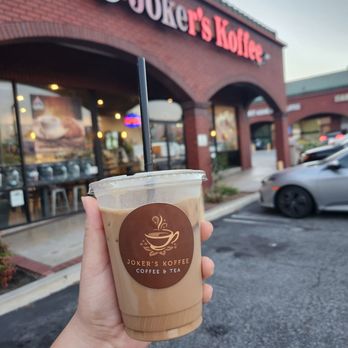 Joker's Koffee - Vietnamese Coffee - A person holds an iced coffee with a Joker's Koffee logo outside the cafe, which has a brick facade and large windows. - Coffee & Tea Near Me - Arcadia, California