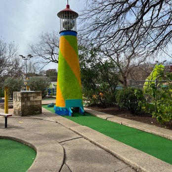 Peter Pan Mini-Golf - A mini golf course features a lighthouse-shaped obstacle painted in yellow, green, and blue. The lighthouse has a red top with a bird weather vane and a cage-like structure around the light. The course has a green artificial turf pathway and surrounding concrete and landscaped areas with trees and bushes. The background shows a partly cloudy sky and outdoor lighting fixtures. - Mini Golf Near Me - Austin, Texas