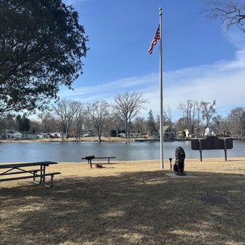 Lions Park - A park features a flagpole with the American flag, a lion statue at its base, and several benches on a grassy area. In the background, there is a lake with houses and leafless trees along the shoreline. The sky is mostly clear with some clouds, indicating a calm day. - Parks Near Me - Fox River Grove, Illinois