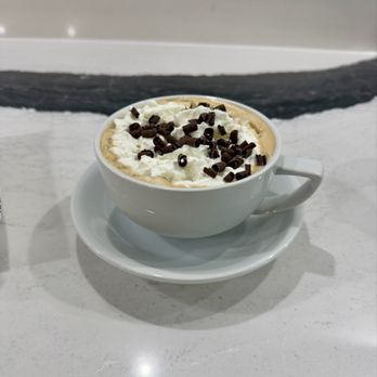 Refuel Coffee - White Christmas - A white ceramic coffee cup filled with a hot beverage, topped with whipped cream and sprinkled with chocolate shavings, is placed on a matching saucer. The cup is on a white marble table with a subtle pattern, and the background features a white wall with a black decorative strip. The setting appears to be in a cafe or coffee shop. - Coffee & Tea Near Me - St. Augustine, Florida