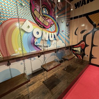 Hurts Donut - Four wooden swings with metal chains are arranged in front of a vibrant, donut-themed mural with rainbow and pop art elements. - Donuts Near Me - Cincinnati, Ohio