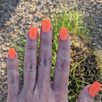 Flashy Nails - A hand with five fingers extended displays bright orange nail polish, set against a gravel and grass background outdoors. - Nail Salons Near Me - Albuquerque, New Mexico