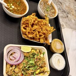 Onion Pakora 8oz Kothu Paratha - The image shows a black tray with Indian food items: a bowl of onion soup, another bowl of a different soup, a serving of onion pakora, and a container of sauce. A separate dish contains a mixed dish garnished with sliced onions, cilantro, and a lime wedge. The tray is placed on a marble surface, and the food appears freshly prepared.