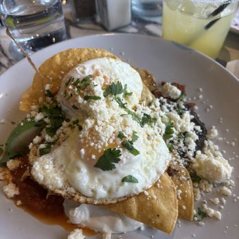 Showroom - Huevos Rancheros - The image shows two poached eggs on a tortilla, topped with cheese, chopped herbs, and sauce, garnished with cilantro. The dish is served on a white plate, with a background including glasses of water, salt and pepper shakers, and a yellow beverage with lime and black straw. - New American Near Me - Frederick, Maryland
