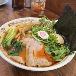 Chili Shoyu Ramen - A bowl of ramen contains sliced pork, bamboo shoots, leafy greens, microgreens, a sheet of nori, and a narutomaki fish cake with a pink swirl. The broth appears rich and flavorful, and the bowl is placed on a wooden table in a restaurant environment. In the background, there are condiment bottles and a glass of iced tea.