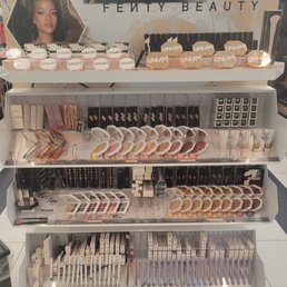A multi-tiered retail display showcases Fenty Beauty makeup products, including creams, powders, and brushes, with branding and promotional images.