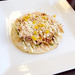 Arepa con Todo - A Colombian-style arepa is presented on a white square plate, topped with shredded chicken, crumbled cheese, yellow corn kernels, and a drizzle of sauce. The arepa appears to be thick and round, with toppings evenly distributed. The background shows a dark surface, emphasizing the dish's presentation.