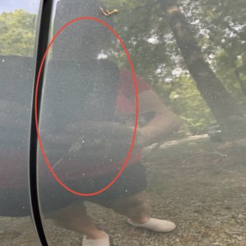 Lund Collision - Clear coat smudging unable to rub off - A photograph of a car's exterior surface shows a smudge or mark on the paint, highlighted by a red oval. The surface is reflective, capturing the reflection of the person taking the photo, along with trees and sky in the background. The person is wearing a red shirt and white shoes, and the ground is covered with small gravel or stones. - Auto Detailing Near Me - Bridgewater, Massachusetts
