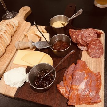 Stem & Stone - The image shows a wooden serving board with an assortment of sliced cured meats, including salami and other charcuterie, along with slices of cheese. Small metal bowls contain sauces or jams, and there is a dollop of cream cheese or soft cheese on the side. Bread slices are arranged on a separate wooden board in the background. The setting appears to be a restaurant or bar table. - New American Near Me - Bastrop, Texas
