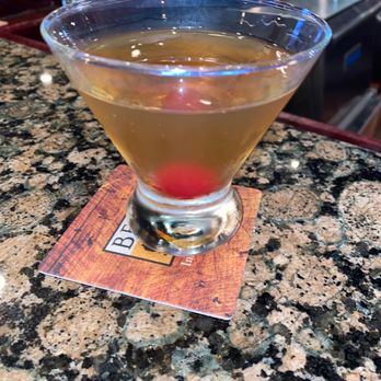 Oscar's Alehouse - Orange Manhattan..... - A cocktail in a martini glass with a cherry at the bottom, placed on a bar counter with a coaster. - New American Near Me - Eldersburg, Maryland