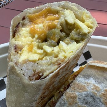 Ursula - My delicious Breakfast burrito - A breakfast burrito with a flour tortilla exterior, cut open to reveal scrambled eggs, melted cheese, green chilies, and other fillings. The burrito is placed in a white takeout container with black-and-white checkered paper lining. The background shows a reddish-brown surface, possibly a table or outdoor seating area. - New Mexican Cuisine Near Me - Brooklyn, New York