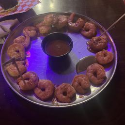 Cajun Shrimp - A round metal tray contains multiple fried shrimp arranged in a circle, with a small cup of dipping sauce in the center and a lemon wedge on the side. The tray is placed on a dark wooden table, with some food items and a checkered paper in the background, indicating a restaurant environment.