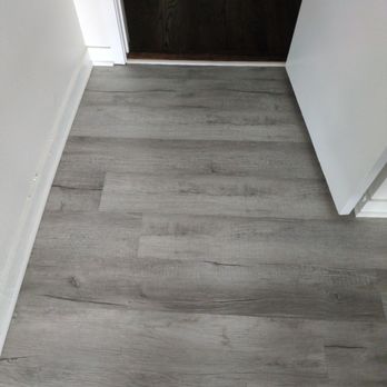 Royal Home Flooring - Flooring installation bedroom - The photo depicts a flooring transition area where light gray laminate flooring meets dark hardwood flooring at a doorway. The laminate planks have a wood grain pattern and are installed parallel to the wall, while the dark hardwood flooring appears to be a different material and color. The transition is clean, with a small threshold strip or molding separating the two types of flooring. - Flooring Near Me - Bridgeview, Illinois