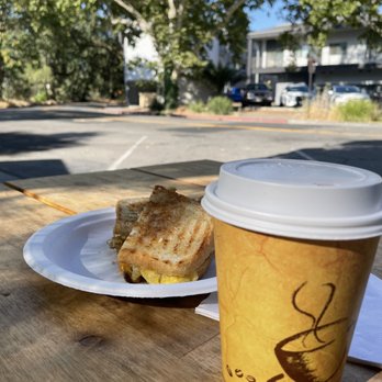 Three Ladies Cafe - small latte  Mushroom, Egg and Cheese Panini - A toasted sandwich on a paper plate and a hot drink in a paper cup are placed on a wooden table outside, with trees and buildings in the background. - Juice Bars & Smoothies Near Me - Davis, California