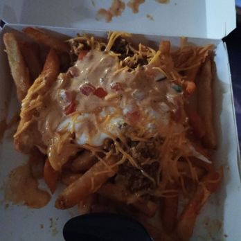 Taco Bell - Jalapeno Ranch Nacho Fries - A food container holds a serving of fries topped with melted cheese, sour cream, diced tomatoes, and other ingredients. The fries are partially visible beneath the toppings, and a black spoon is placed inside the box. The container appears to be a typical fast-food or takeout box, with some sauce and toppings spilled around the edges. - Fast Food Near Me - Espanola, New Mexico