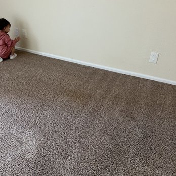 Motif - stain - A young girl wearing a pink jacket and white shoes is seated on a brown textured carpet, facing a plain off-white wall with multiple electrical outlets. The room appears to be a residential or indoor space with minimal visible furniture or decor. - Apartments Near Me - Phoenix, Arizona