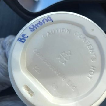 Zeb's Coffeehouse - A white disposable coffee cup with a plastic lid is shown, with the handwritten phrase 'BC strong' in blue ink on the lid. The cup appears to be held by a person, with part of their clothing visible. The background includes a vehicle interior, suggesting the photo was taken inside a car. - Coffee & Tea Near Me - Eudora, Kansas