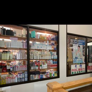 Angel Beauty - Inside the entrance of the building - A storefront window of Angel Beauty in Boston displays multiple shelves filled with skincare, cosmetic, and beauty products. The products are organized in rows, with packaging in various colors and sizes. The window has a sign with the business name and a poster featuring a woman holding beauty products. The scene includes a wooden railing in the foreground, indicating the store's exterior view. - Cosmetics & Beauty Supply Near Me - Boston, Massachusetts