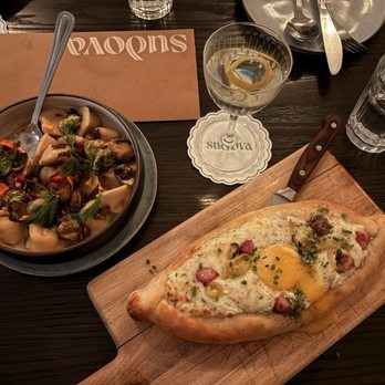 Sudova * Eastern European Kitchen - khachapuri special with kielbasa sausage and egg with butternut squash dumplings - The image shows a khachapuri with egg and kielbasa on a wooden board, alongside a bowl of dumplings with vegetables and garnishes. - Modern European Near Me - Cincinnati, Ohio