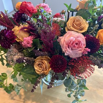 Fiori Flowers - A large, vibrant bouquet of mixed flowers is arranged in a clear glass vase placed on a wooden table. The bouquet includes roses, dahlias, and other blooms in shades of pink, purple, yellow, and red, complemented by green foliage and trailing greenery. The background is neutral, with natural light illuminating the flowers. - Florists Near Me - Boulder, Colorado