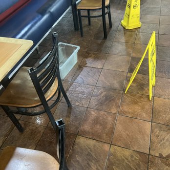 KFC - The image shows a section of a restaurant with a tiled floor that appears wet, as evidenced by the reflections and water marks. Two yellow caution signs are placed on the floor, warning of a wet surface. There are several chairs with wooden seats and black metal frames, some positioned around tables with light-colored wooden tops. A clear plastic container is on the floor near the chairs. The seating area includes a blue cushioned bench along the wall, which has a decorative element. The overall scene suggests ongoing cleaning or recent spillage in the dining area. - Fast Food Near Me - Maricopa, Arizona