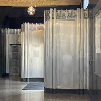 The Basement - First floor of the building, technically above The Basement - The photo depicts an indoor space featuring metallic wall panels with decorative patterns, characteristic of art deco design. An elevator door with ornate detailing is visible on the left side, and the floor is covered with a speckled, polished surface. The ceiling has decorative molding and a hanging spherical light fixture. A window on the right side allows natural light into the space. - Public Markets Near Me - Reno, Nevada
