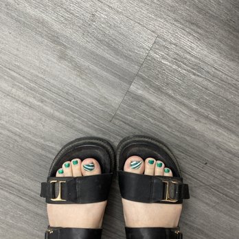 Smiley Nails - Feet in black sandals with gold buckles, showing toenails painted green with white and green striped designs, on a gray wooden floor. - Nail Salons Near Me - Phoenix, Arizona