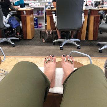 Famous Nails - The image shows a person's feet with gel nails, featuring black and light green nail polish, resting on a towel-covered footrest in a nail salon. The background includes nail polish displays, salon staff working at a counter, and various salon tools and supplies. The person is wearing olive-green pants, and the photo is taken from their perspective, focusing on their feet and the salon environment. - Nail Salons Near Me - Albuquerque, New Mexico