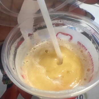 Smoothie King - A yellow smoothie is in a clear plastic cup with a straw, placed on a table. The drink appears frothy and is likely fruit-flavored. - Juice Bars & Smoothies Near Me - Laurel, Maryland