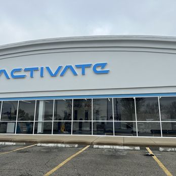Activate Games - The image shows the exterior of Activate, a business with a white curved facade and large glass windows. The blue sign displaying 'ACTIVATE' is prominently positioned on the building. The parking lot in front has marked parking spaces, some of which are occupied. The sky above is overcast, and the building's address, 8600, is visible on the right side of the facade. - Team Building Activities Near Me - Fishers, Indiana