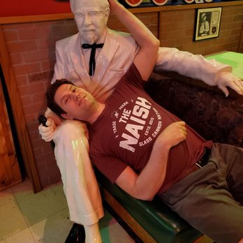 Old School Pizza Malta - True Love - A man is reclining on a bench in a restaurant or bar, next to a large statue of a man in a tuxedo, with a brick wall and framed pictures in the background. - Pizza Near Me - Malta, Illinois