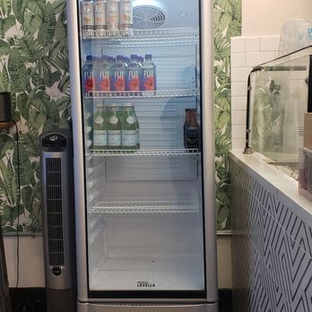 Acai bar  - Drink frig - A glass-front refrigerator containing canned and bottled beverages, placed next to a fan and a patterned counter, with leafy wallpaper in the background. - Acai Bowls Near Me - Pasadena, California