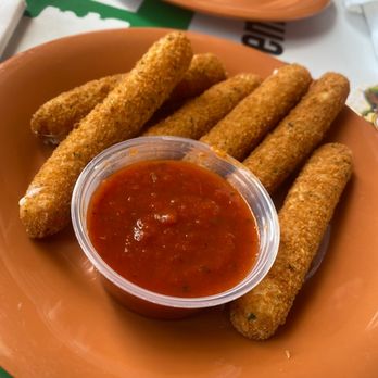 Mulligans Pub and Grill - A plate containing several breaded and fried mozzarella sticks arranged around a small cup of marinara sauce is shown. The mozzarella sticks are golden brown and crispy. The plate is orange, and the background includes another empty plate, napkins, and tableware, indicating a casual dining setting at Mulligans Pub and Grill, which offers appetizers like mozzarella sticks and a variety of other menu items. - Pubs Near Me - Avon, Ohio