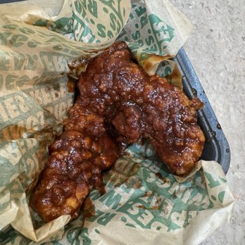 Wingstop - Sweet bbq blaze tenders - Two glazed fried chicken pieces are placed on a black tray, wrapped in paper with green text, likely from a fast-food restaurant. - Chicken Wings Near Me - Fairfax, Virginia