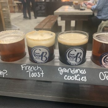 Crue Brew Brewery - Flight of four - 21 pumpkins, French toast, grandmas cookies, and old rusty wagon - Four craft beers are arranged on a wooden flight board at Crue Brew Brewery. Each beer is in a branded glass with the brewery's logo, and the board has handwritten labels indicating the beer names: '21 Pump.', 'French Toast', 'Grandma's Cookies', and 'O2W'. The background shows a casual brewery setting with patrons seated at tables and a person in a blue shirt working on a device. - Breweries Near Me - Raynham, Massachusetts
