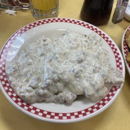 Biscuits and Gravy - A bowl of biscuits and gravy with a thick, creamy white sauce is on a yellow table at Ze's Diner. The bowl has a red and white checkered rim. In the background, there is a glass of orange juice, a dark syrup bottle, and other dining items. The setting suggests a casual American diner environment.