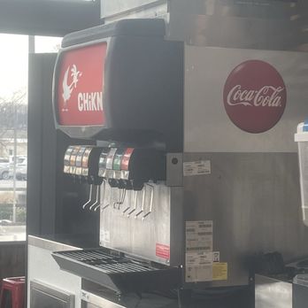 CHiKN - Soda and Lemonade Tea stand - A soda fountain machine featuring Coca-Cola branding and multiple beverage flavor options, with a drip tray below the dispensers. The machine is placed on a stainless steel counter near a window, with a plastic container and other equipment nearby. The setting appears to be inside a restaurant or cafe, with natural light coming through the window. - Chicken Shop Near Me - Crafton, Pennsylvania