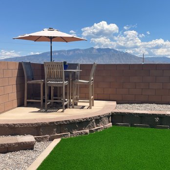 AMG Landscape - Curved section of the block wall, with a colored concrete top section for our patio table. - A patio with a high brick wall, outdoor table, chairs, and a large umbrella, with mountains and clouds in the background. - Irrigation Near Me