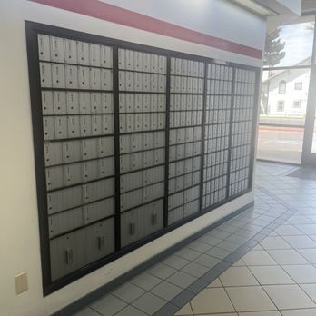 US Post Office - The image shows a large wall-mounted mailbox unit with multiple individual metal mailboxes arranged in a grid pattern. The mailboxes are housed within a black frame and are located inside a building, likely a post office or apartment lobby. The floor features tiled flooring with a pattern of square tiles, and there is a glass door and window to the right, allowing natural light and a view of the outside, including a building and some trees. - Post Offices Near Me - Gilbert, Arizona