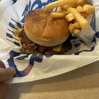 Culver's - My dinner which is The Culver's Bacon Deluxe Double - A burger with a soft bun, melted cheese, and a beef patty is presented in branded paper. Crinkle-cut fries are placed on top of the burger, with additional fries in a paper container. The meal is served on a tray at a fast-food restaurant, with a hand holding the tray visible in the image. - Fast Food Near Me - Marion, Indiana