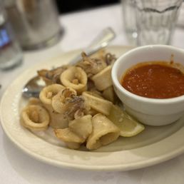 Fried Calamari - Fried calamari rings are arranged on a white oval plate, accompanied by a small bowl of marinara sauce and a lemon wedge. The dish is placed on a white tablecloth with glasses and utensils visible in the background, indicating a restaurant setting.