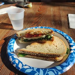 Veggie monster - A sandwich with visible vegetables, cheese, and bread is placed on a blue and white patterned paper plate on a wooden table. A clear plastic cup filled with water is positioned behind the plate. The setting appears to be a casual dining area, with a person partially visible in the background, along with a smartphone and a napkin on the table.